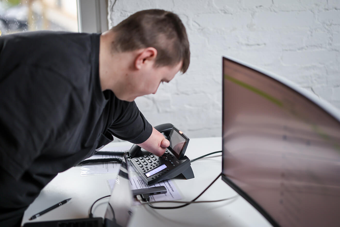 Yannick Boos, Büromanager | © Rupert Oberhäuser Ein junger Mann mit verkürzten Armen beugt sich über den Schreibtisch. Mit seinem Armstumpf tippt er auf die Tasten des Telefons.