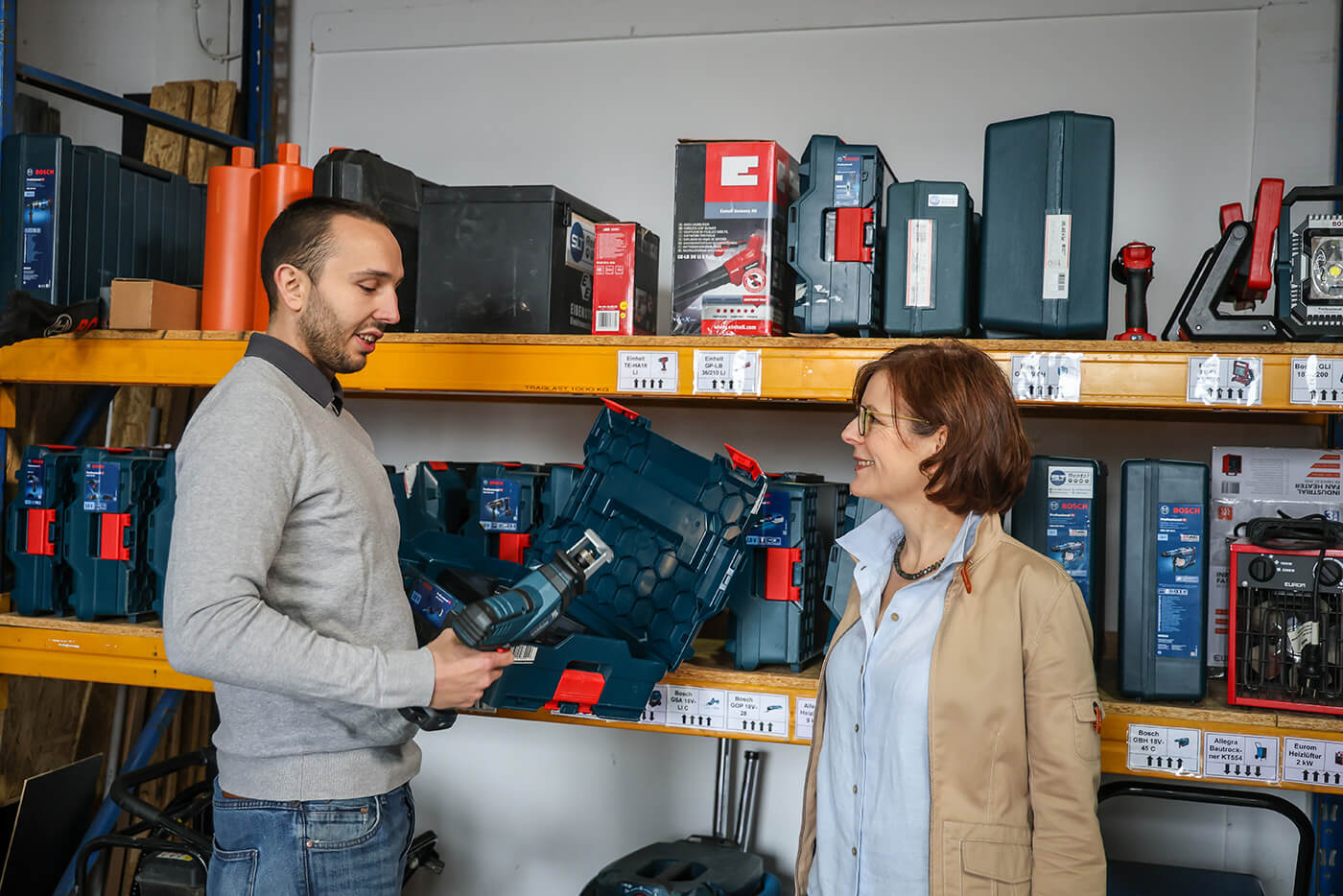 Ein Mann und eine Frau stehen in einem Lager vor einem Regal mit verschiedenen Kartons. Der Mann hält eine Verpackung in den Händen, die er der Frau zeigt.