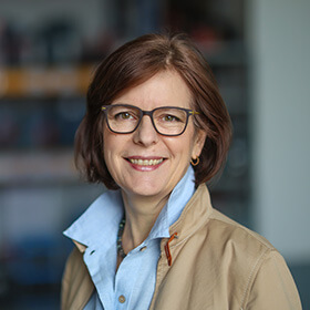 Eine Frau mit blauer Bluse, beigefarbener Jacke und Brille schaut lächelnd in die Kamera.