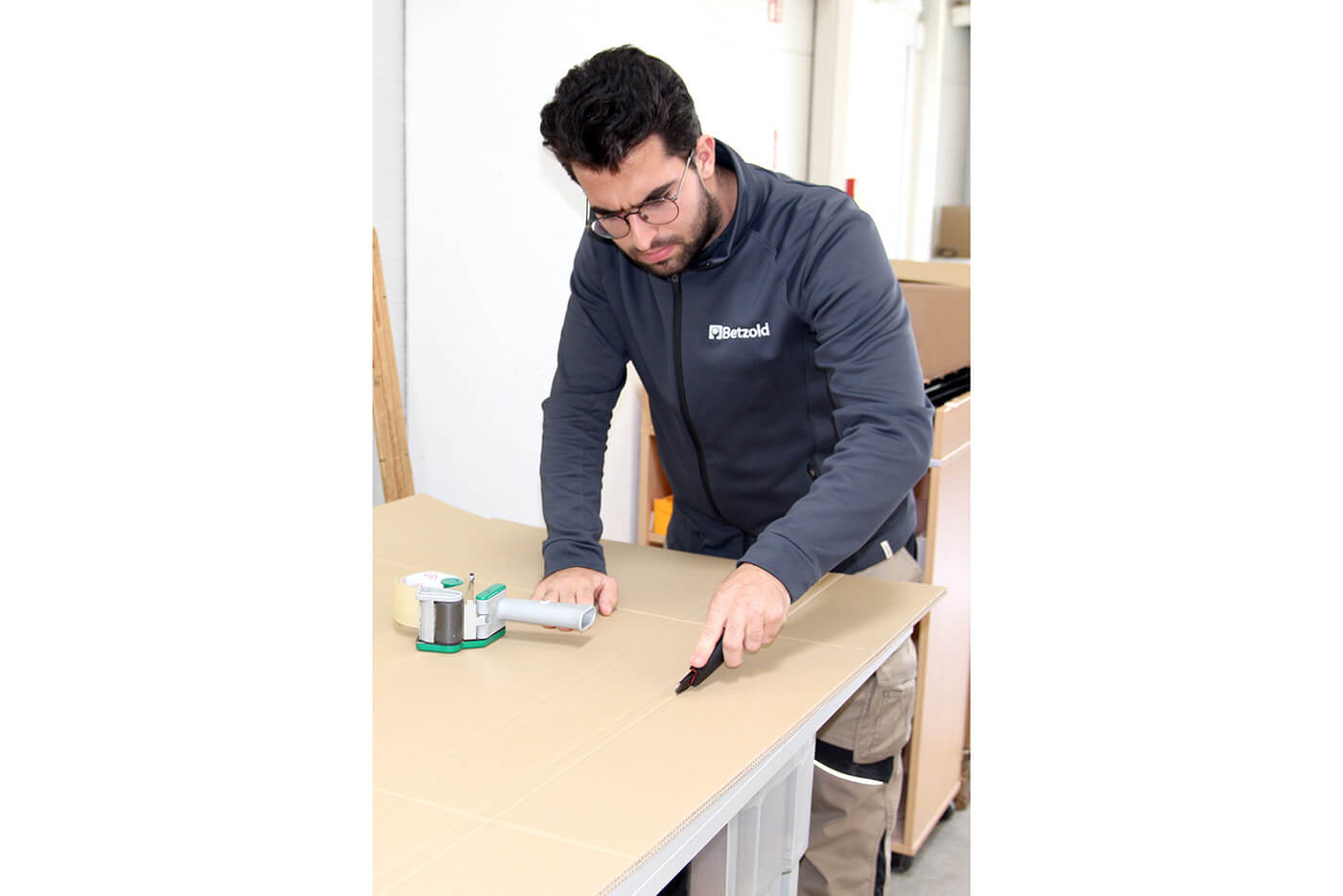 Isa-Onur Sarioglu schneidet an einem Werktisch einen großen Karton mit einem Cuttermesser zu.