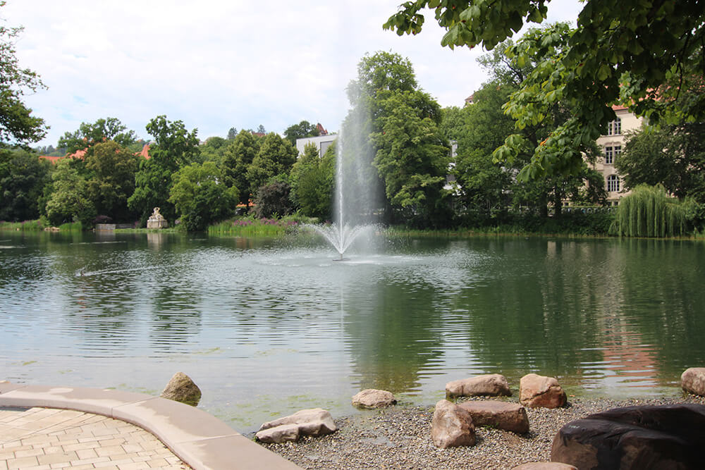 Ein kleiner See mit einer Wasserfontäne. Am hinteren Ufer stehen viele Bäume, am vorderen Ufer sieht man noch den Rand einer Terrasse, daneben beginnt ein kleiner Kiesstrand, der in den See führt.