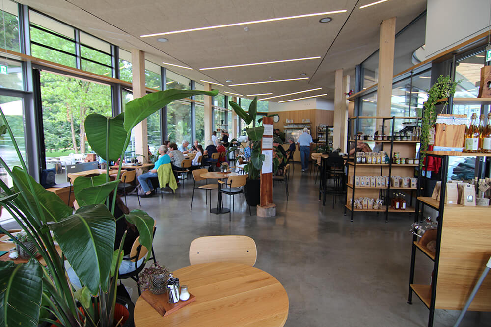Der helle Café-Raum mit runden Holztischen, links vorne im Bild ist eine große Grünpflanze zu sehen, dahinter die große Glasfront. Rechts sind Holzregale mit verschiedenen Produkten wie Teepackungen oder Weinflaschen.