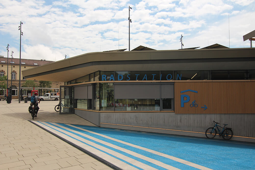 Im Vordergrund ist die Abfahrt zur Fahrradtiefgarage zu sehen, die nach rechts unten führt. Die Abfahrt ist in hellem blau gestrichen. Sie grenzt direkt an die Rückseite der Fahrradstation, die in einem eingeschossigen Flachdachgebäude mit Holzfassade untergebracht ist.