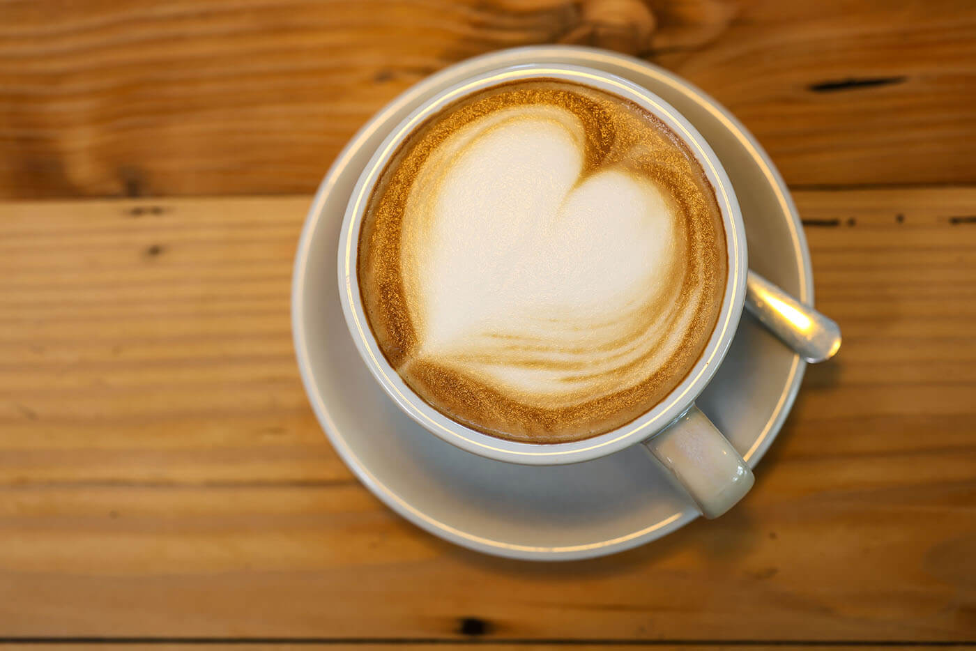 Eine weiße Kaffeetasse mit einem Cappuccino steht auf einem Naturholztisch. Der Milchschaum in der Kaffeetasse ist zu einem Herzen geformt. 