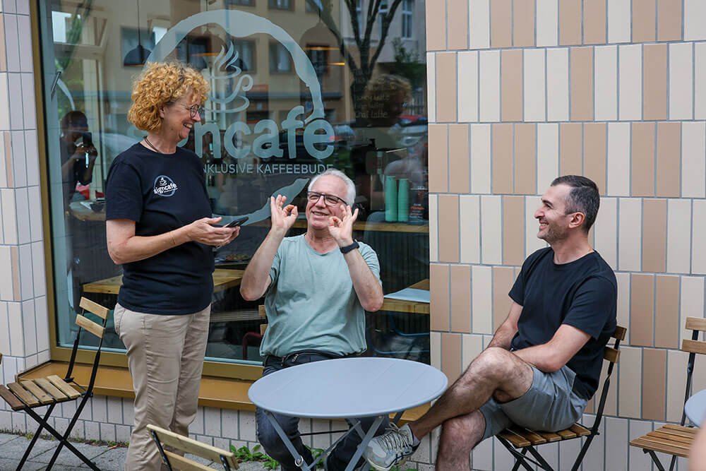 Das signcafe für Gehörlose, Schwerhörige und Hörende | © Rupert Oberhäuser Zwei Männer sitzen draußen an einem runden Tisch vor einem Schaufenster mit der Aufschrift signcafe. Eine Frau mit rotblonden Locken bedient. Die Kommunikation läuft über Zeichensprache.