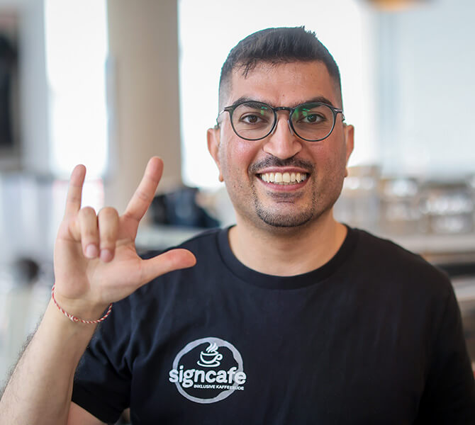 Mitarbeiter Zeredesit Omar | © Rupert Oberhäuser Ein junger Mann in einem schwarzen T-Shirt und dem weißen Aufdruck "signcafe. Inklusive Kaffeebude"formt mit der rechten Hand die Gebärde für "I love you".