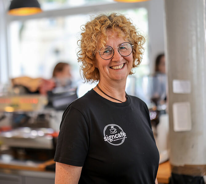 signcafe-Inhaberin Angela Herbig | © Rupert Oberhäuser Eine Frau mittleren Alters mit rotblonden Locken und schwarzem T-Shirt lächelt direkt in die Kamera. Auf dem T-Shirt steht "signcafe. Inklusive Kaffeebude"