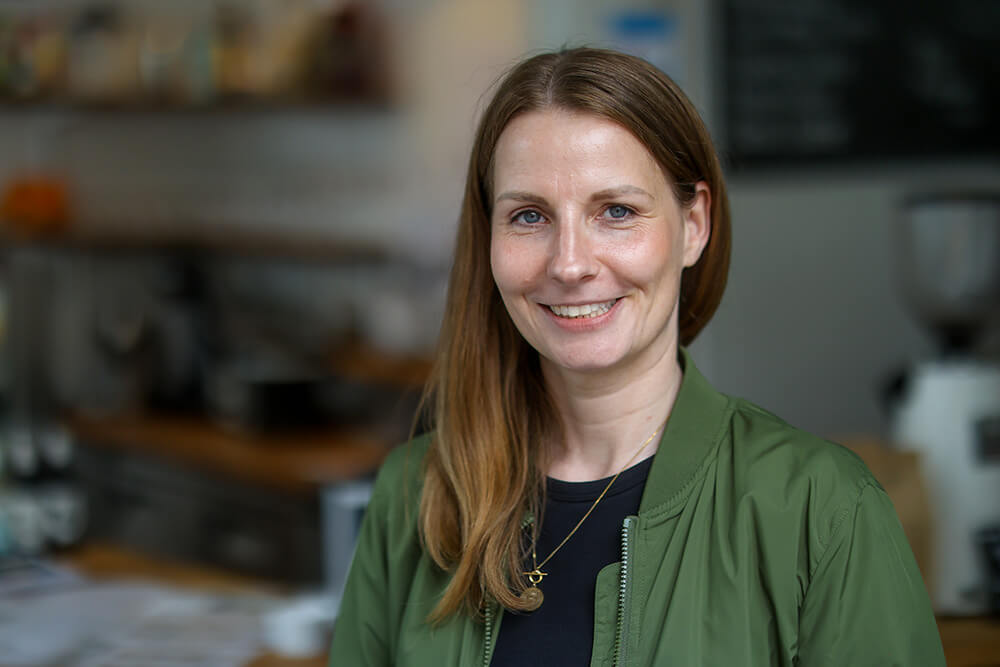 LVR-Mitarbeiterin Simone Zimmer | © Rupert Oberhäuser Eine junge Frau mit langen Haaren und grüner Blouson-Jacke steht lächelnd in einem Café.