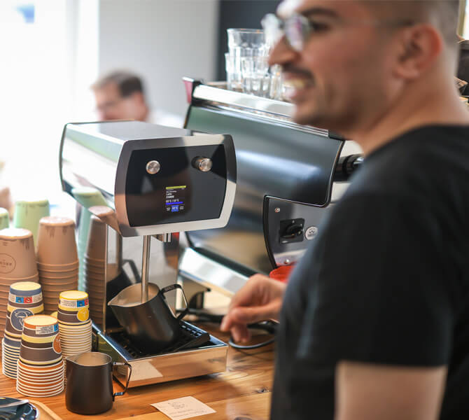 Zeredesit Omar am barrierefreien Milchschäumer | © Rupert Oberhäuser Ein junger Mann im schwarzen T-Shirt steht in einem Café vor einem elektrischen Milchaufschäumer.