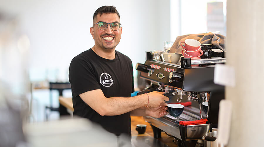 Ein junger gehörloser Mann steht in einem hellen, freundlichen Café an der Kaffeemaschine. Lächelnd zapft er einen Espresso.