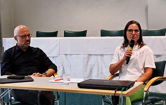 Ein Mann und eine Frau sitzen bei einer Tagung hinter einem Tisch, auf dem Tisch stehen Laptops und liegen Papiere. Die Frau hält ein Mikrophon in der Hand.