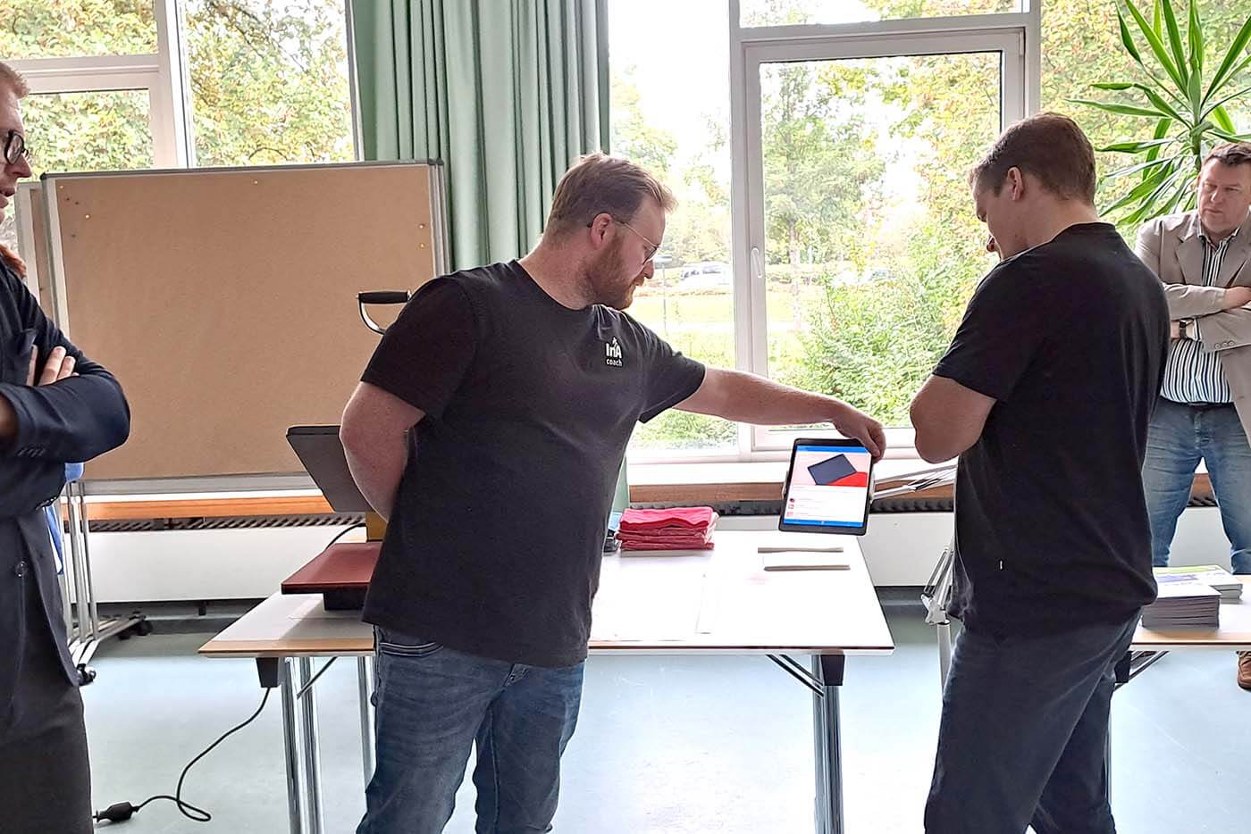 Ein Mann steht in einem Seminarraum vor einem Tisch und zeigt einem anderen Mann an einem Tablet, wie die neue App funktioniert