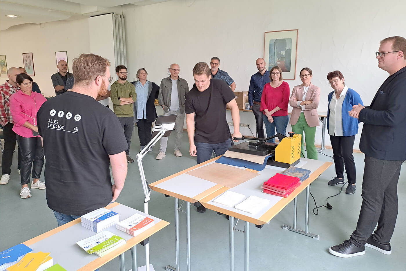 In einem Seminarraum stehen viele Menschen locker um einen Tisch gruppiert. An dem Tisch ist eine Halterung für ein Tablet befestigt. Zweijunge Männer stehen vor dem Tablet und demonstrieren die Anwendung der App.