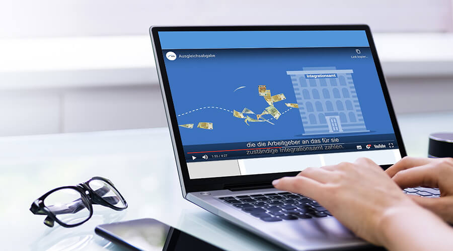 Neue Stufe der Ausgleichsabgabe | ©Andrey_Popov/Shutterstock Auf einem Laptop-Monitor ist ein Ausschnitt eines Erklärfilms zur Ausgleichsabgabe zu sehen. In der Szene fliegen Geldscheine in ein Gebäude, das die Inklusions- und Integrationsämter symbolisiert.