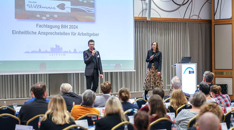 Netzwerkreffe der EAA-Fachberaterinnen und -berater | © BIH/ Rupert Oberhäuser BIH-Vorstandsvorsitzender Christoph Beyer steht auf einer großen Bühne vor einem vollbesetzen Saal und spricht in ein Hand-Mikrophon. Im Hintergrund ist eine Präsentation an die Wand geworfen.