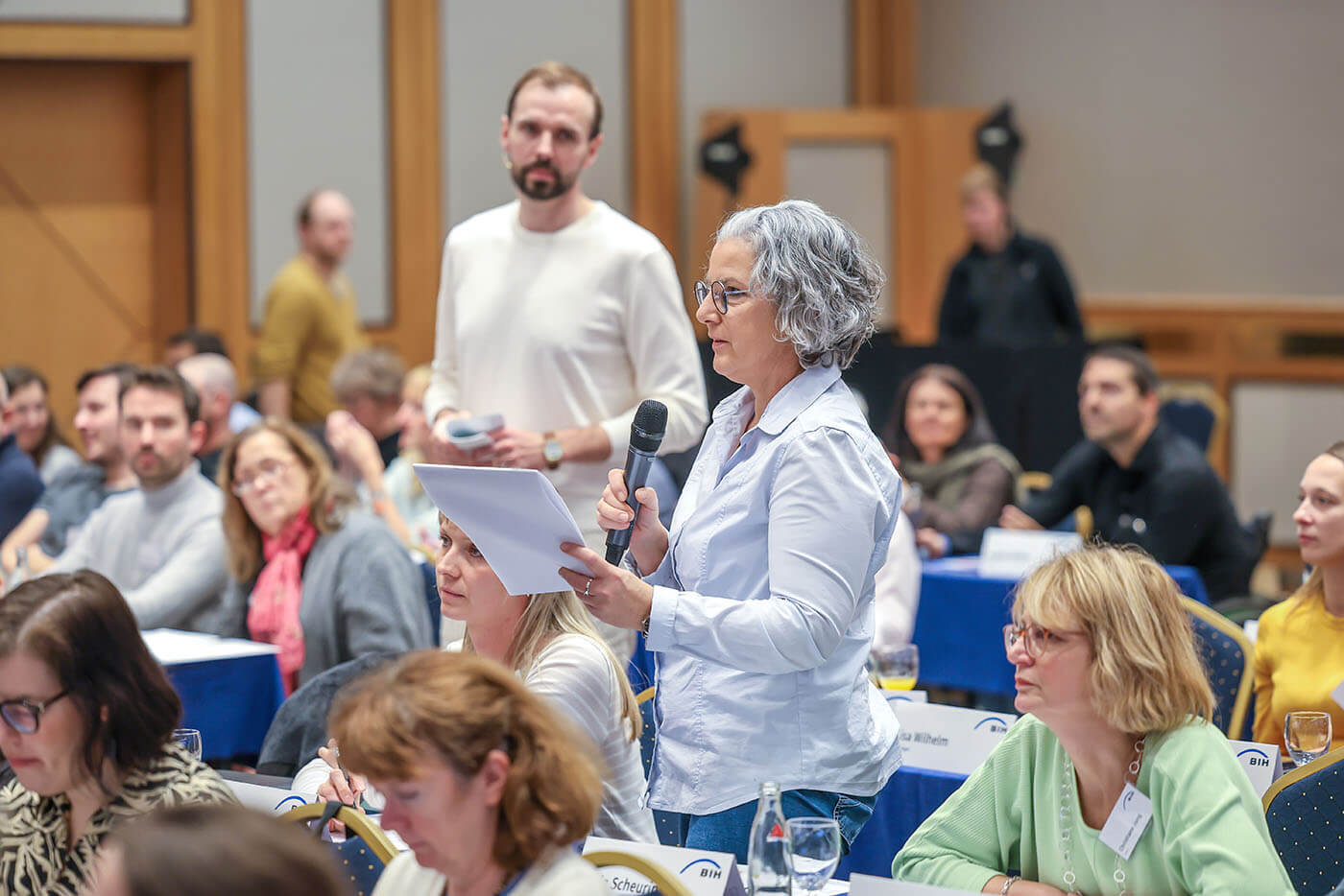 EAA-Netzwerktreffen Diskussionsrunde l ©BIH/Rupert Oberhäuser Eine Frau steht zwischen vielen Menschen, die an Tischen sitzen. Sie hält ein Manuskript in den Händen.