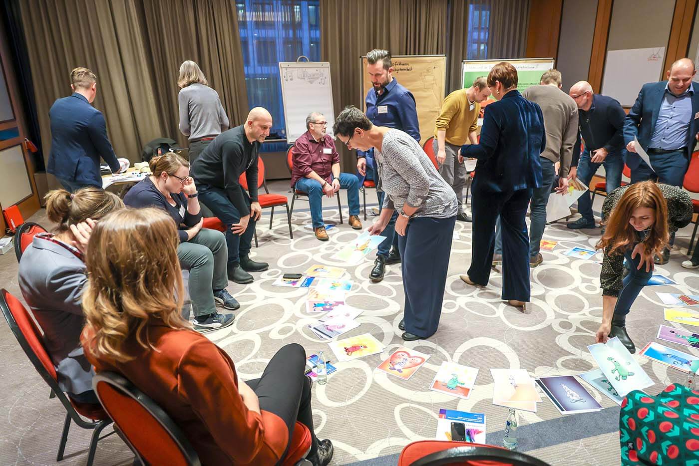 Teilnehmer des Workshops sitzen in einem Stuhlkreis. Mehrere Teilnehmer stehen in der Mitte des Kreises, während weitere Teilnehmer im Hintergrund die Notizen auf den dort aufgestellten Flip-Charts lesen.