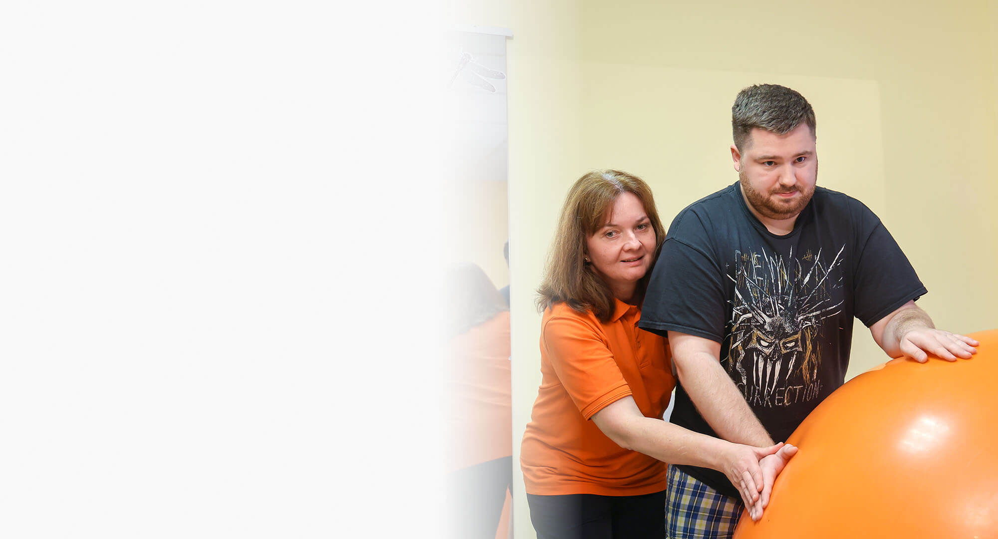 Physiotherapie mit großem Gymnastikball l © BIH/Rupert Oberhäuser Eine Physiotherapeutin behandelt einen jungen Mann, der sich auf einen großen Gymnastikball abstützt. Die Therapeutin steht hinter ihm.