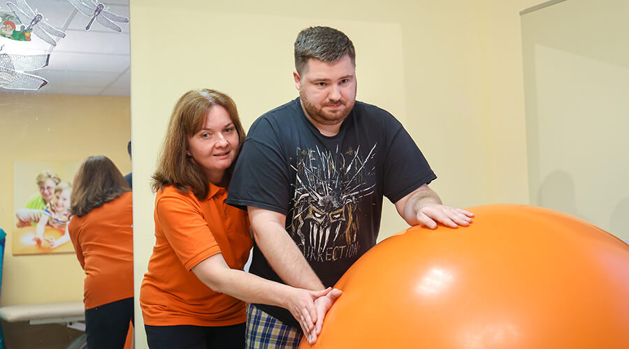 Physiotherapie mit großem Gymnastikball l © BIH/Rupert Oberhäuser Eine Physiotherapeutin behandelt einen jungen Mann, der sich auf einen großen Gymnastikball abstützt. Die Therapeutin steht hinter ihm.