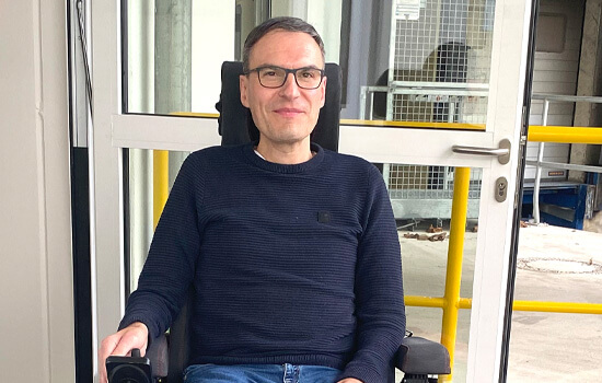 Thomas Schäffers sitzt in seinem Rollstuhl. Der Rollstuhl steht vor der geöffneten Eingangstür seines Arbeitgebers. 