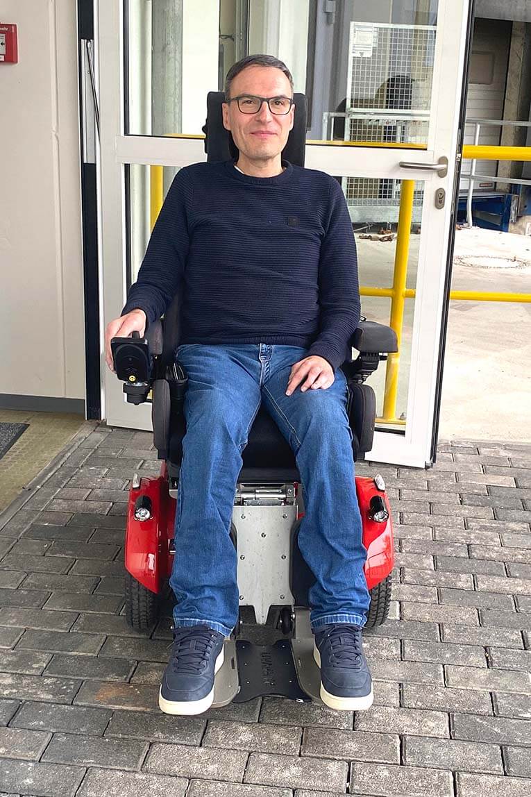 Thomas Schäffers sitzt in seinem Rollstuhl. Der Rollstuhl steht vor der geöffneten Eingangstür seines Arbeitgebers. 