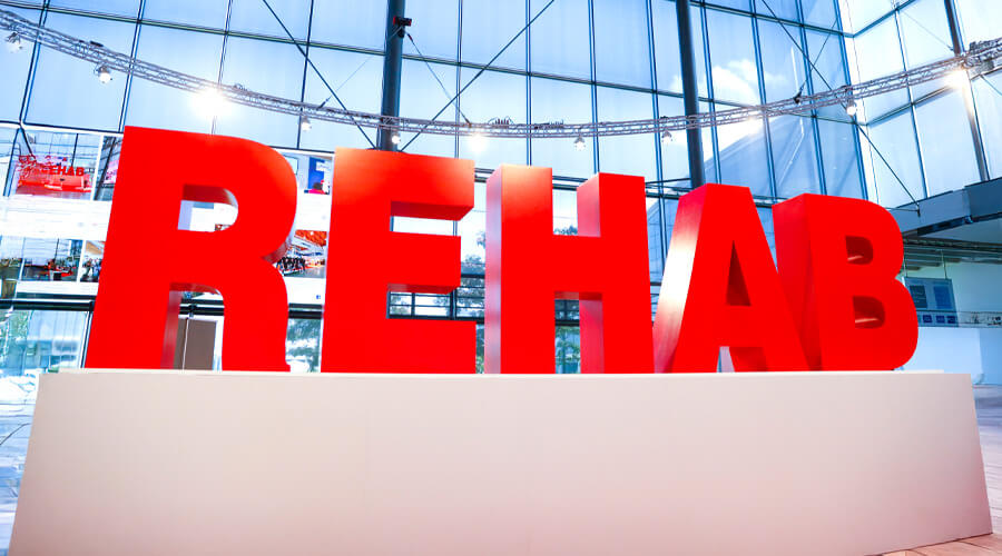 In roten großen Buchstaben steht das Wort "REHAB" auf einem weißen Podest vor einer verspiegelten Wand.