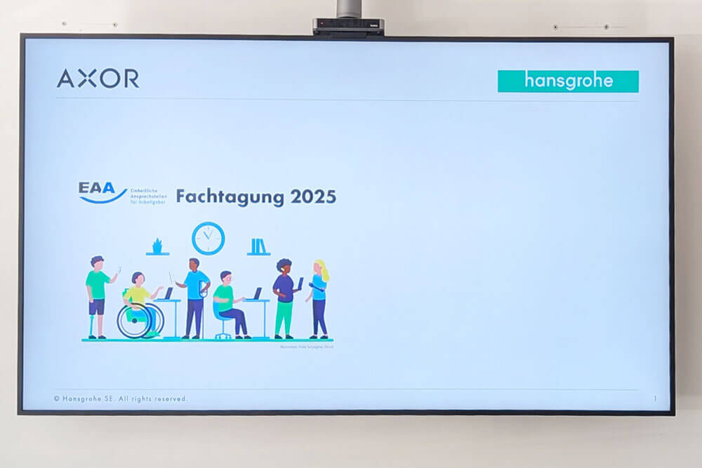 Aufnahme eines großen Bildschirms, der an der Wand hängt und auf dem die Präsentation zum Inklusionskonzept von Hansgrohe zu sehen ist.
