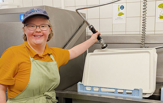 Frederike Gabel steht an der Industriespülmaschine. In der Hand hält sie eine Wasserbrause, mit der sie das Geschirr abspült. 