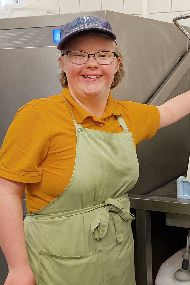 Frederike Gabel steht an der Industriespülmaschine. In der Hand hält sie eine Wasserbrause, mit der sie das Geschirr abspült. 