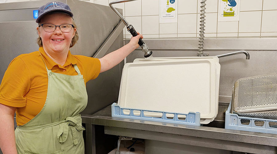 Frederike Gabel an der Spülmaschine l © Monika Kleusch Frederike Gabel steht an der Industriespülmaschine. In der Hand hält sie eine Wasserbrause, mit der sie das Geschirr abspült.