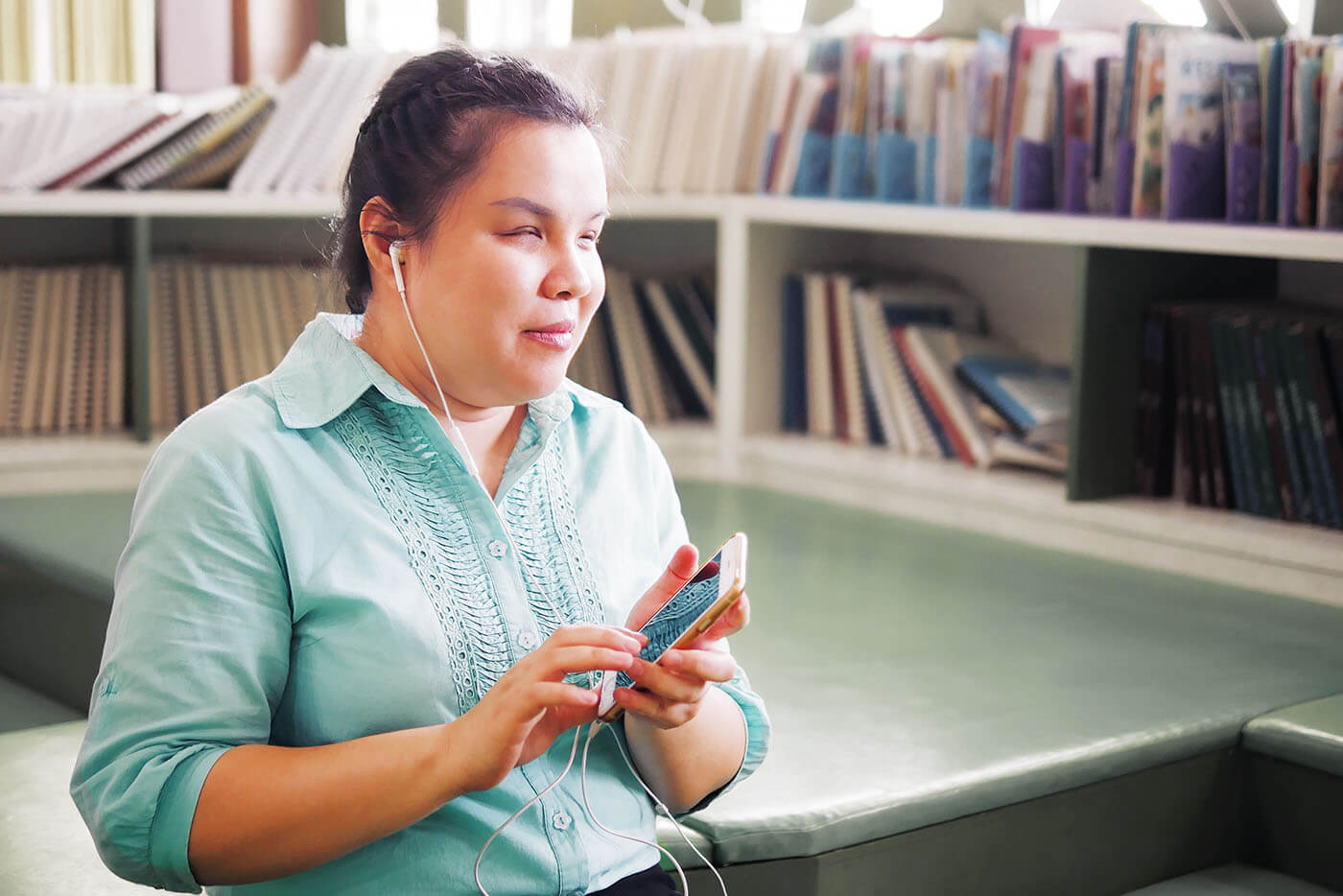 Blinde Frau mit Handy und Kopfhörer l © 2023 Chansom Pantip/Shutterstock Eine blinde Frau sitzt in einer öffentlichen Bibliothek. In den Händen hält sie eine Smartphone. In den Ohren hat sie In-Ear-Kopfhörer, die an das Handy angeschlossen sind