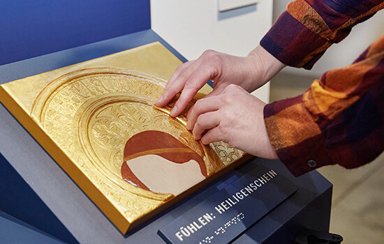 Nahaufnahme einer goldenen Heiligenschein-Plastik, die von zwei Händen ertastet wird.