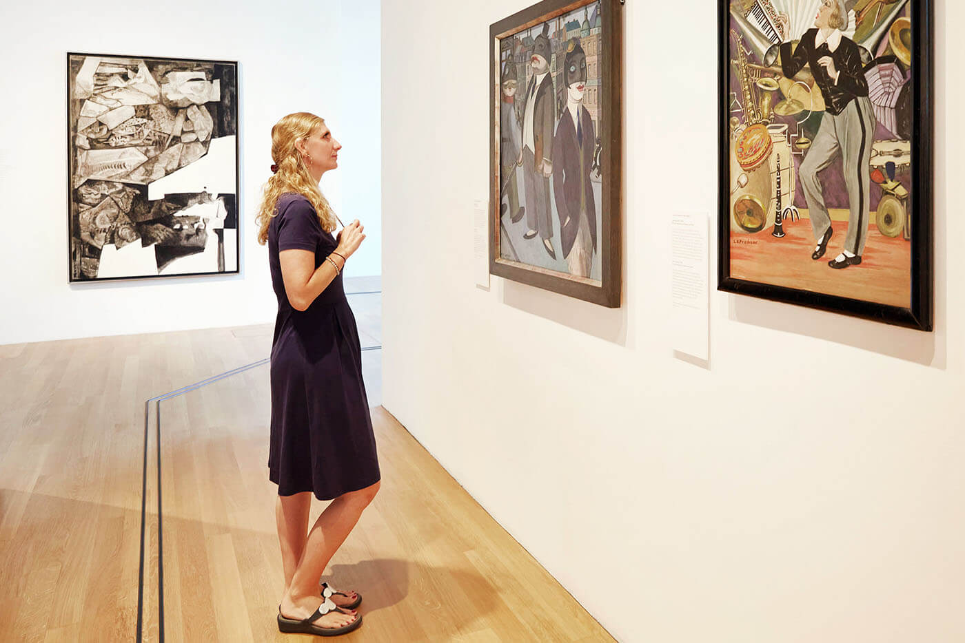 Ein Frau betrachtet Bilder in einem Museum Bilder an einer Wand, auf dem Boden ist ein Tast-Leitsystem für Menschen mit Sehbehinderung zu sehen.