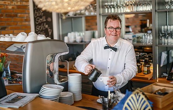 Arne Krüger-Brörken lächelt in die Kamera während er hinter dem Tresen eine Kaffee zubereitet.