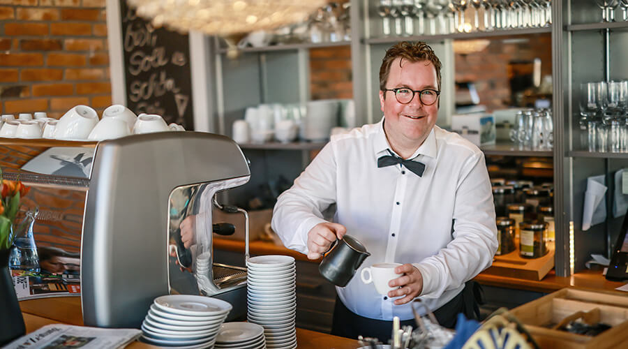 Arne Krüger-Brörken an der Kaffeemaschine l © Rupert Oberhäuser Arne Krüger-Brörken lächelt in die Kamera während er hinter dem Tresen eine Kaffee zubereitet.