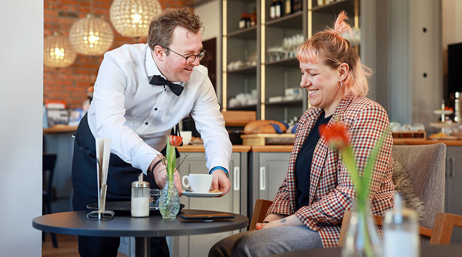 Arne Krüger-Brörken mit Gast l © Rupert Oberhäuser Arne Krüger-Brörken stellt eine Kaffeetasse auf einen Cafétisch vor einem Gast ab.