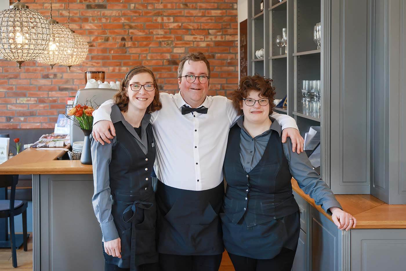 Hotel Villa Sterne Arne Krüger-Brörken mit zwei Kolleginnen l © Rupert Oberhäuser Im Café steht Arne Krüger-Brörken Arm in Arm mit zwei Kolleginnen und lächelt in die Kamera.