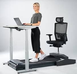 Eine Frau steht auf einem Laufband am Schreibtisch, hinter ihr ist ein Stuhl auf einer Schiene über dem Laufband angebracht