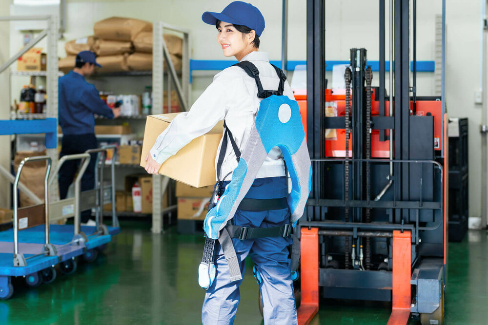 Exoskelette in der Arbeitswelt l © 2023 metamorworks/Shutterstock Eine junge Frau in Arbeitskleidung und einem Exoskelett am Rücken trägt ein großes Pakte in eine Lagerhalle.