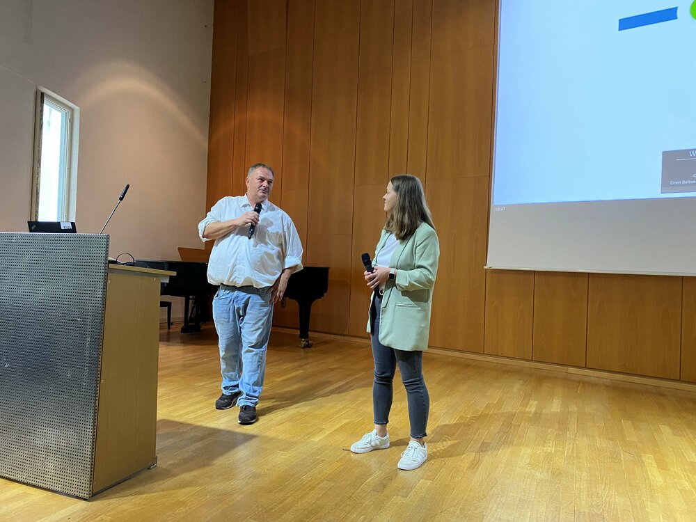Vor einer Leinwand, auf der eine Powerpoint-Präsentation zu sehen ist, stehen zwei Redner mit Mikrophonen.