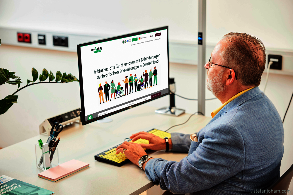 Bildschirm zeigt die Startseite von myAbility.jobs. Darauf ist zu lesen “Inklusive Jobs für Menschen mit Behinderungen und chronischen Erkrankungen in Deutschland“.  Ein Mann mit grauem Haar, Bart und Brille nutzt eine speziell kontrastreiche Tastatur.
