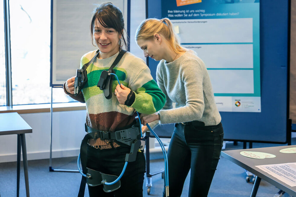 TBD-Fachtagung | Foto: Rupert Oberhäuser Eine junge Frau probiert an einem Messestand ein Exoskellette an. Eine andere junge Frau hilft ihr beim Anpassen.